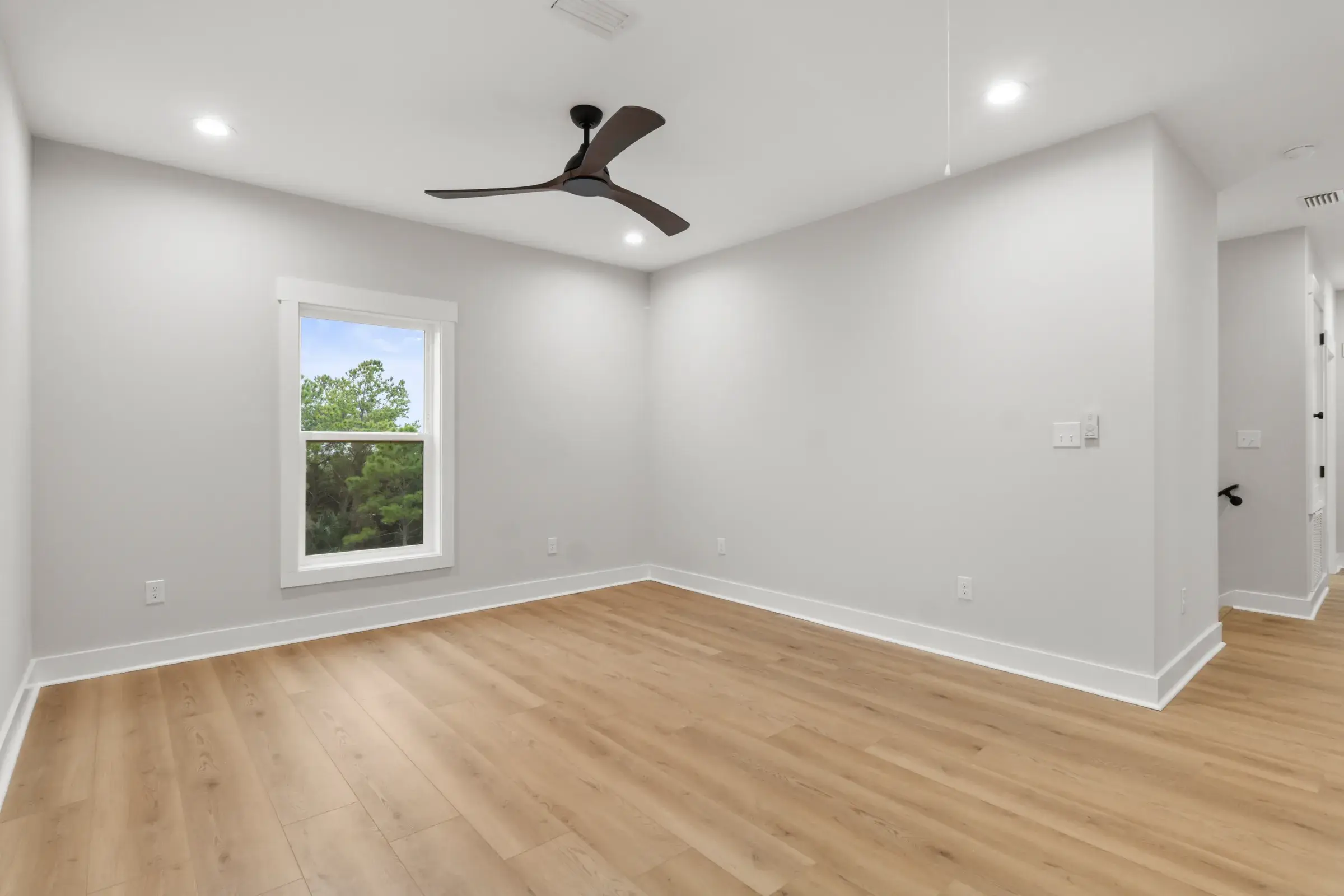 Bedroom with a ceiling fan, a window looking onto trees, and an open hallway on the right.