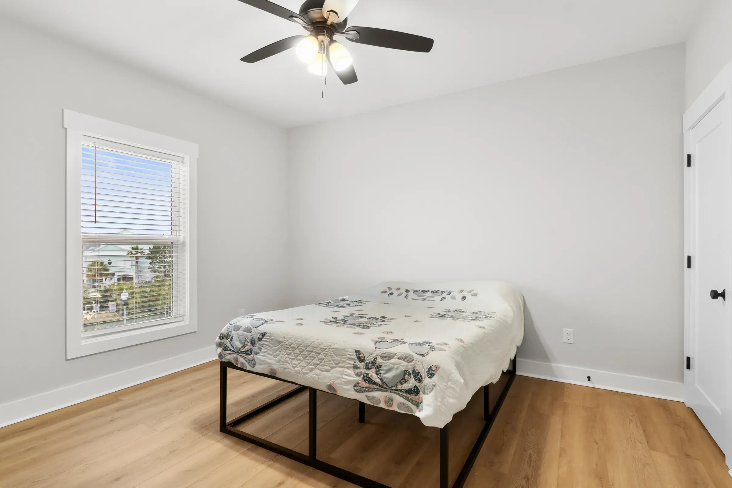 Bright bedroom with a bed on a black metal frame, ceiling fan, window with blinds, and wood-look flooring.