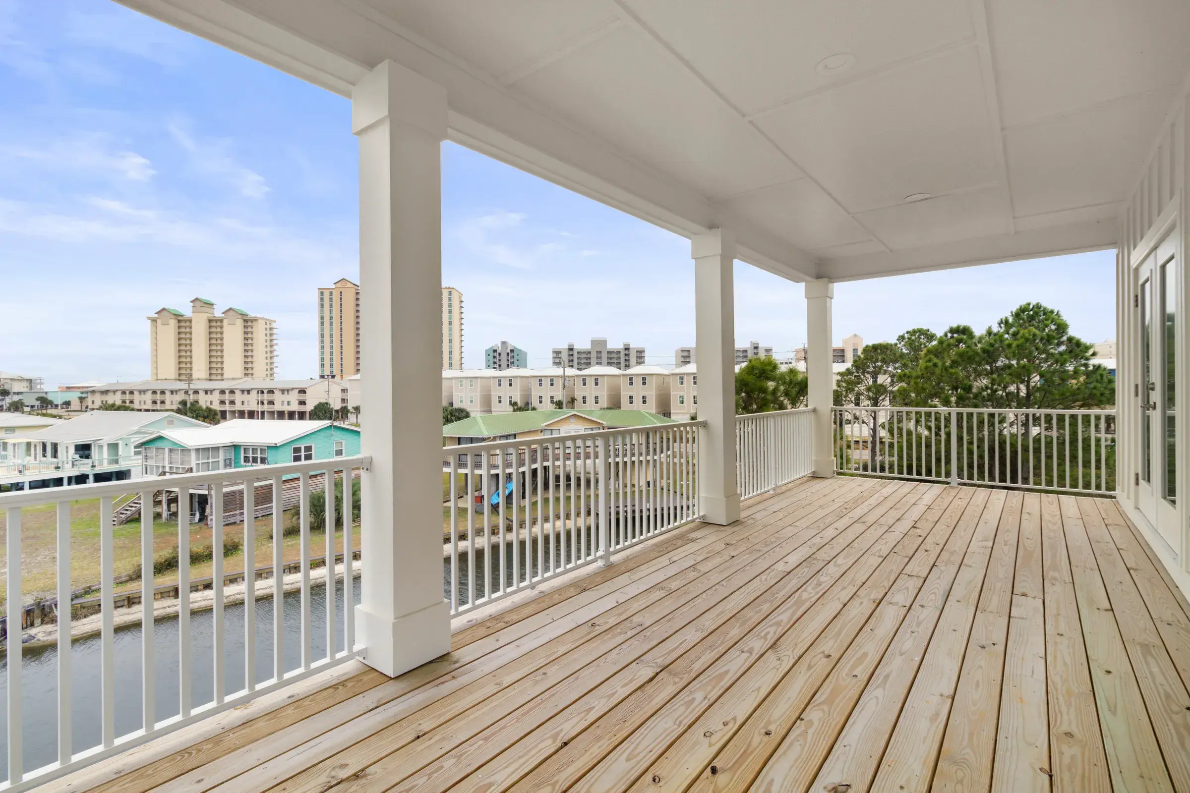 Wide covered wood porch with white columns and railing overlooking the lagoon and neighboring homes.