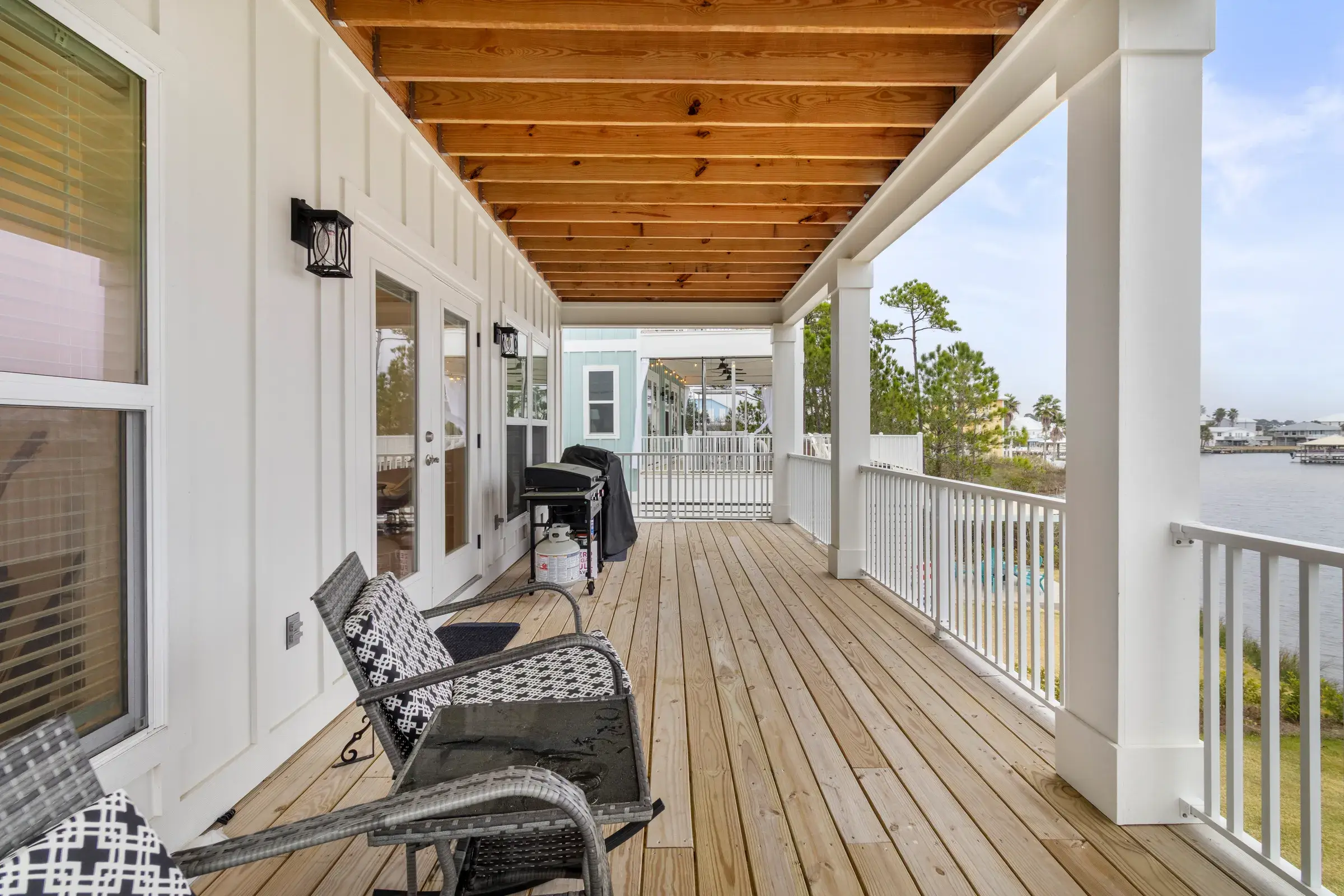 Long covered wood porch with outdoor chairs, a grill at the far end and railings facing the water.