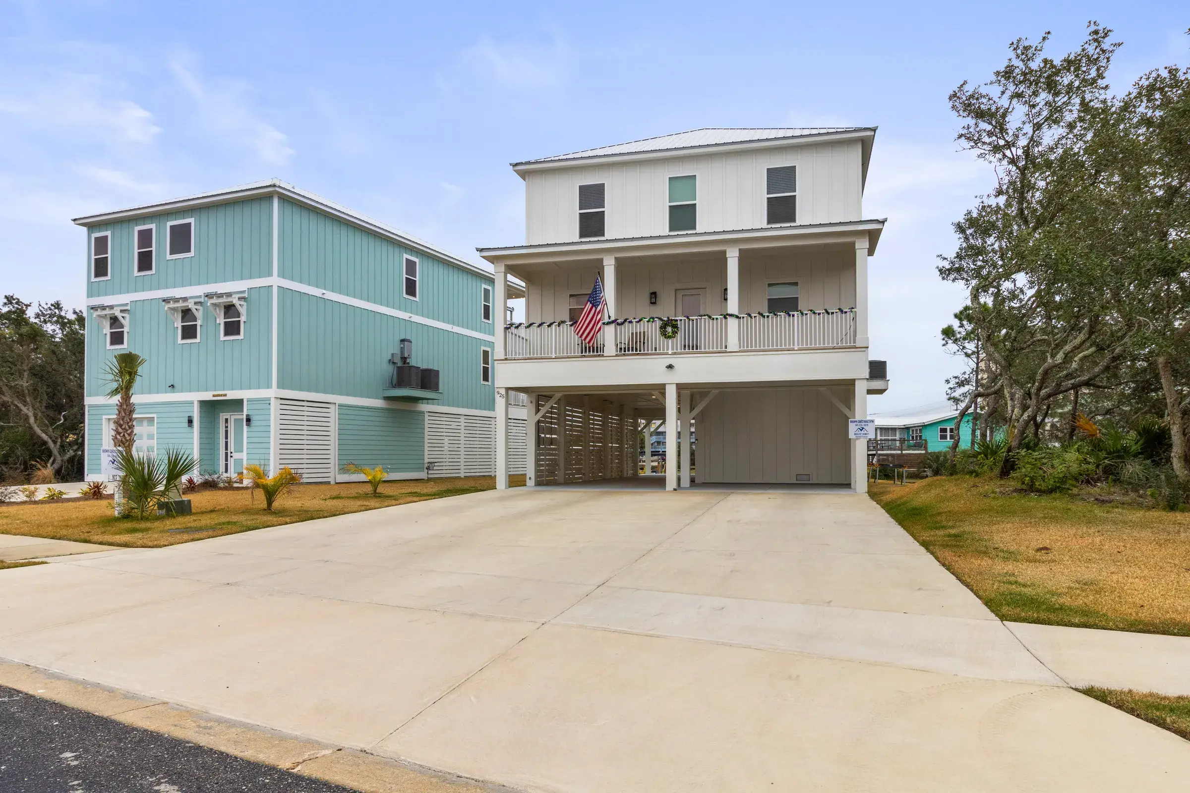 Two-story white home on raised pilings with a covered front porch, flag and wide concrete driveway.