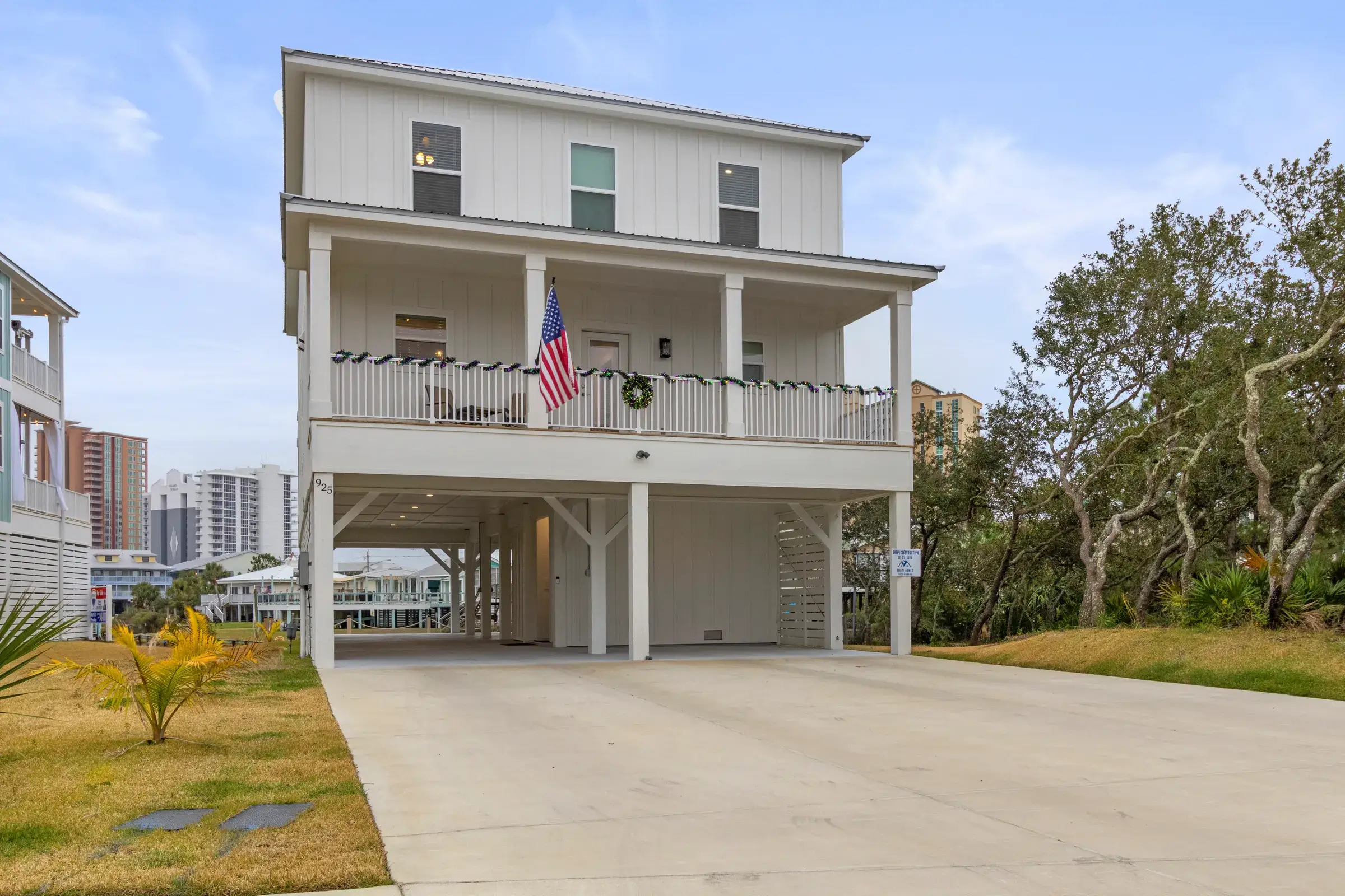 Two-story white home on pilings with a covered upper porch, flag, and open carport over a wide concrete driveway.