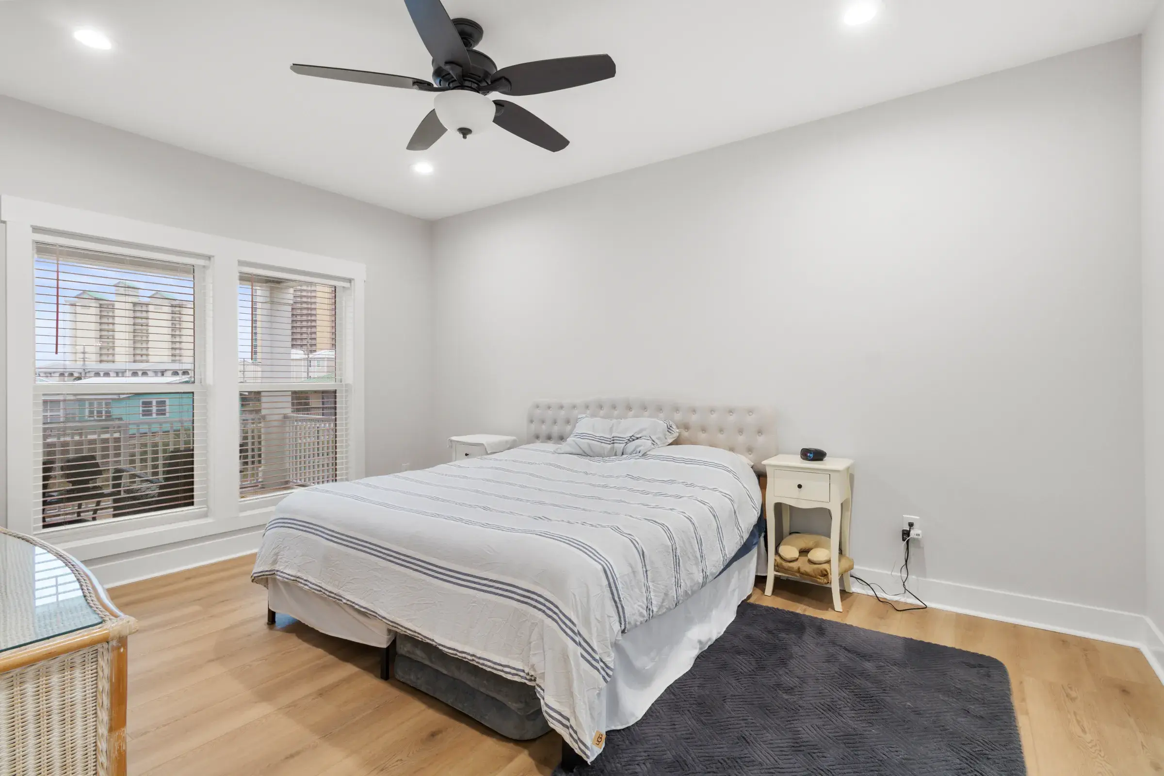 Furnished bedroom with a queen bed, two nightstands, ceiling fan, and double windows with blinds.