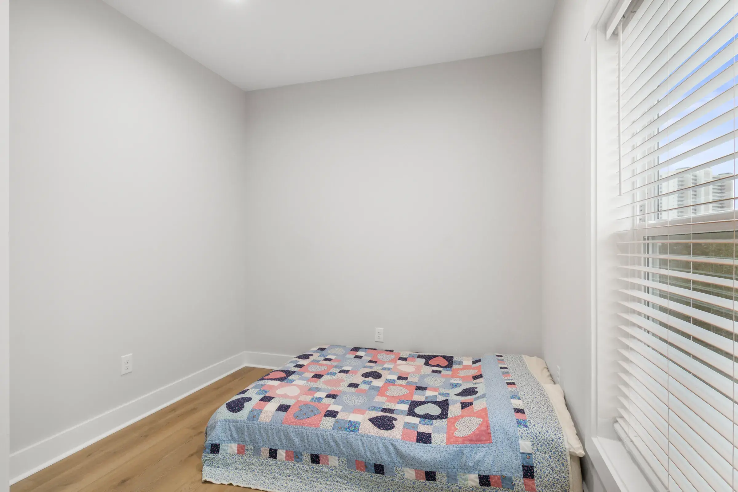 Bedroom with a quilted bed, large window with blinds, and light wood-look flooring.