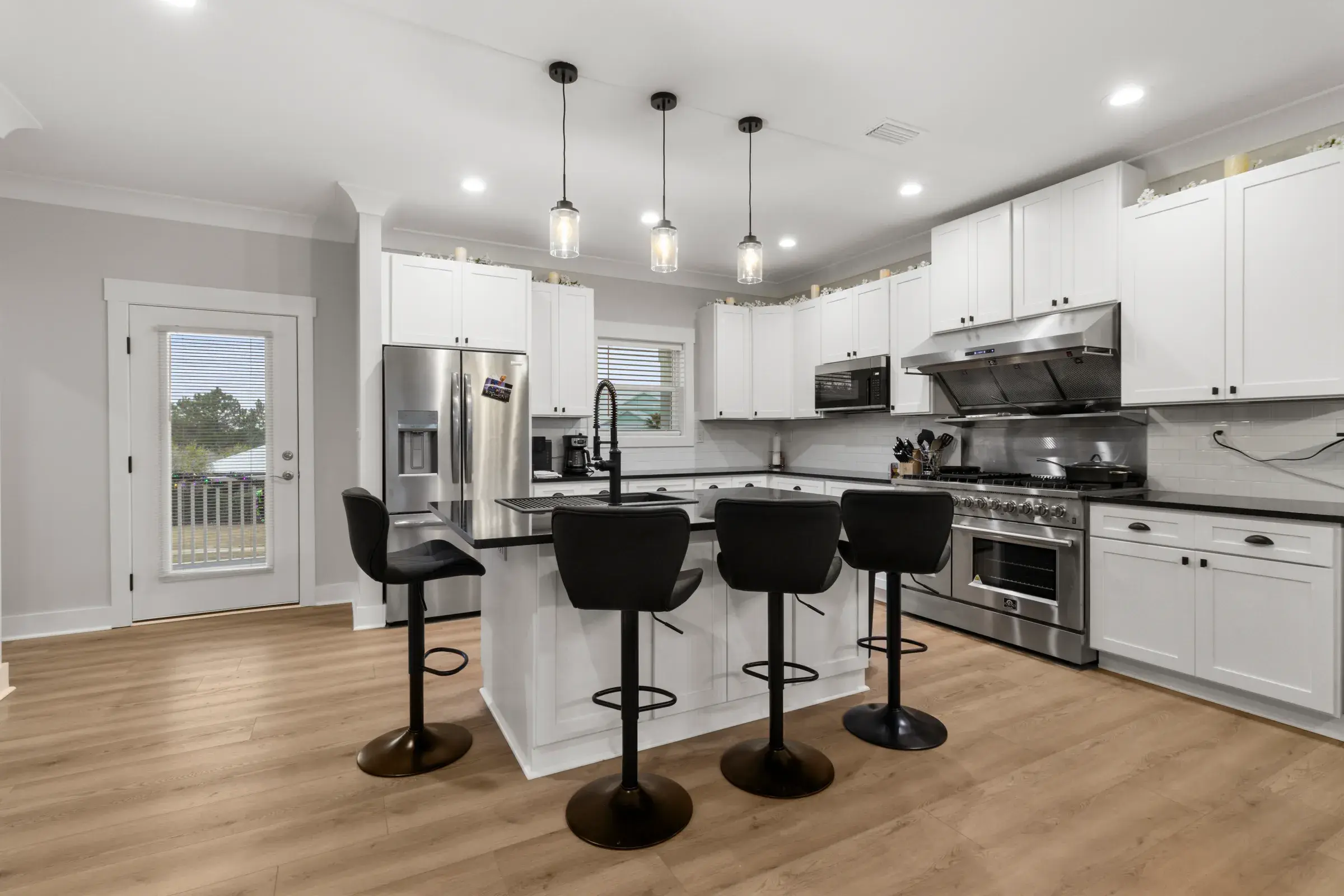 White kitchen with dark granite island, black barstools, pendant lights, stainless refrigerator and a gas range under a hood.