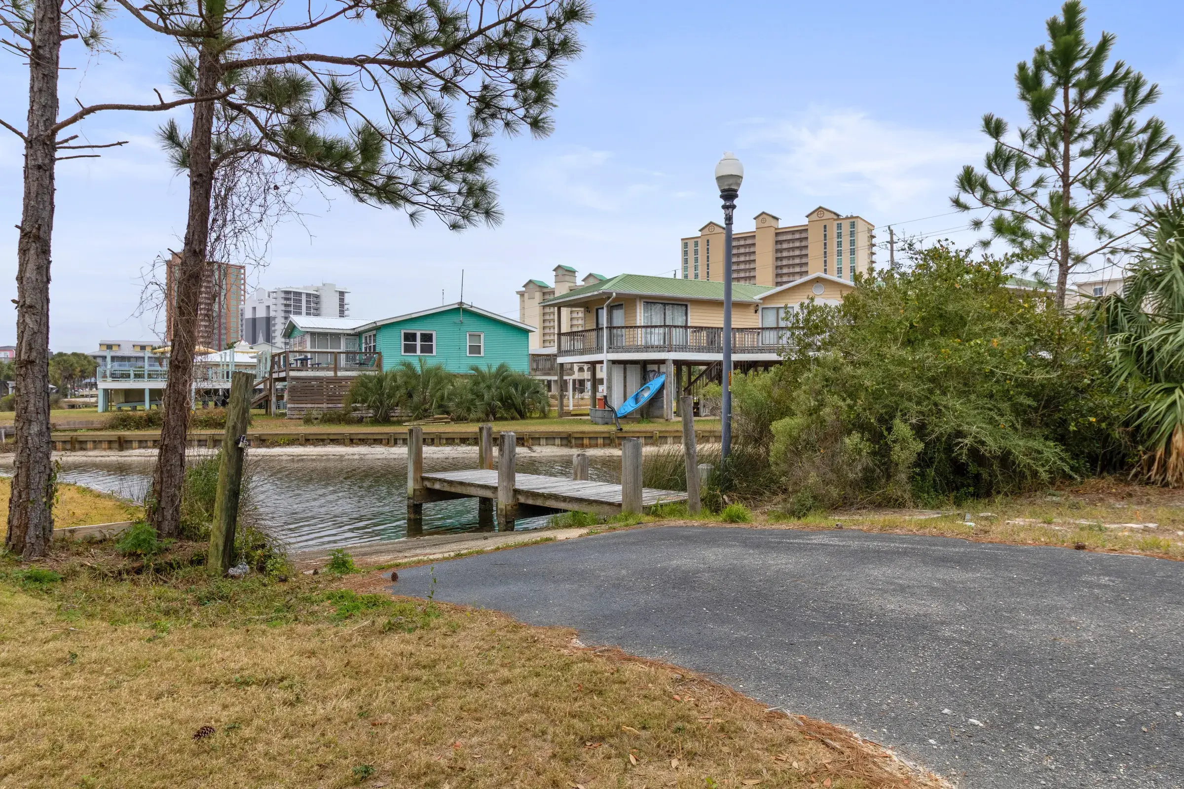 Small wooden dock on the lagoon next to a paved ramp, with waterfront homes and pine trees beyond.