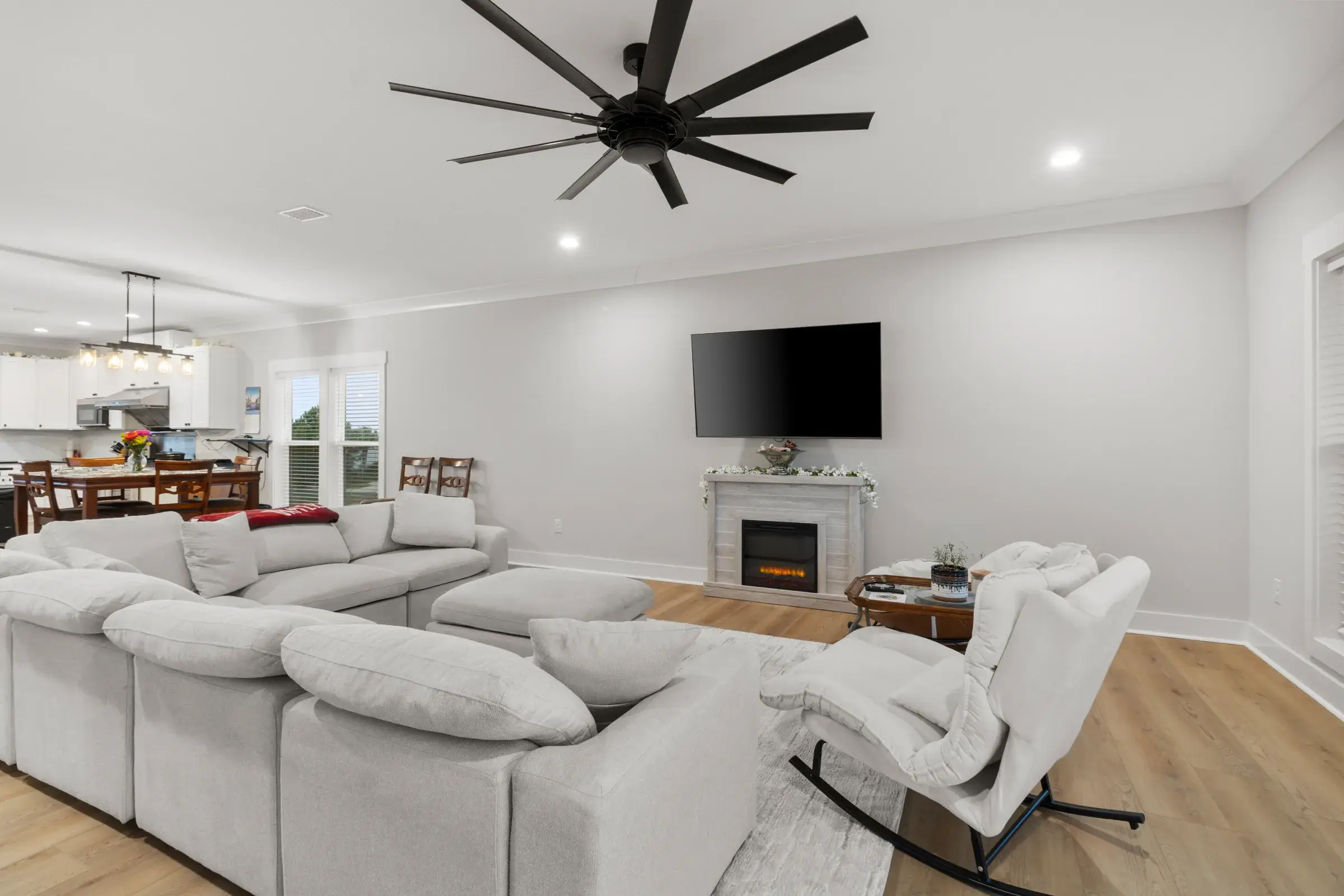 Living room with a gray sectional, large ceiling fan, and fireplace, with the dining table and kitchen beyond.