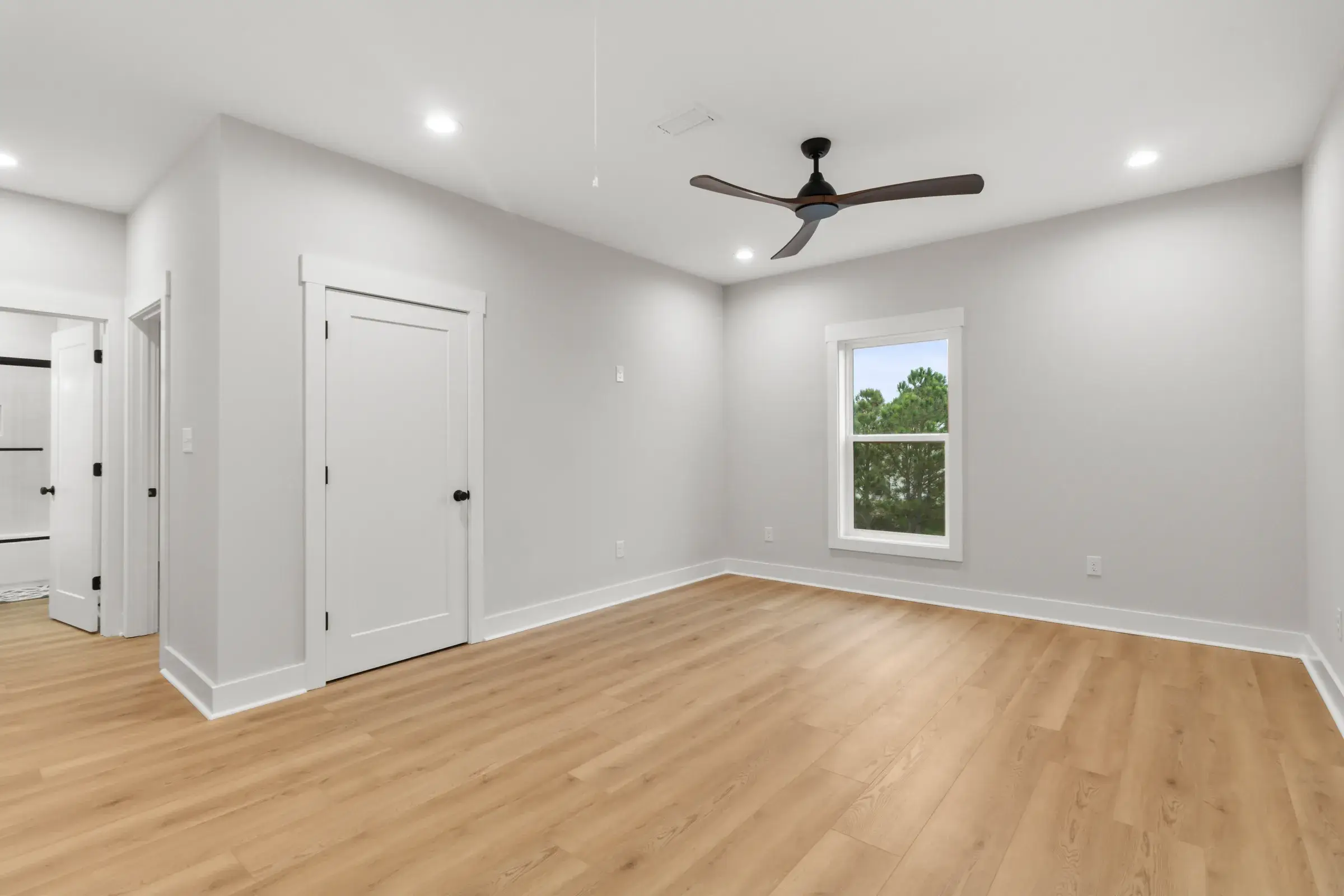 Large bedroom with a dark ceiling fan, recessed lights, closet door, window, and light wood-look floors.